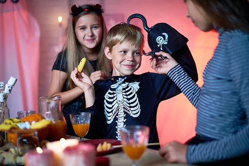 Girl showing toy scorpion to smiling cute boy in skeleton costume with hook instead of hand eating cookie at Halloween party, cheerful pretty girl behind him