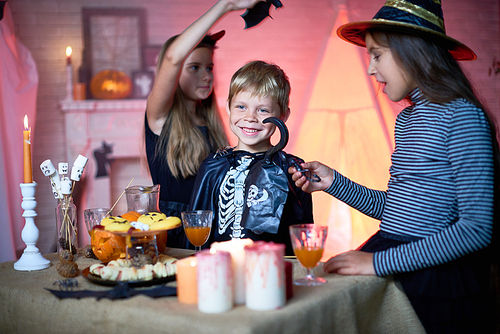 Portrait of children wearing Halloween costumes playing in decorated room during party, standing by table with food and sweets