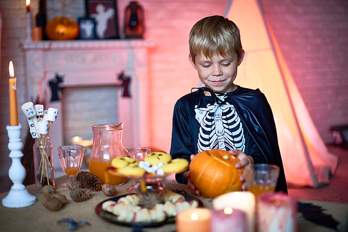 Happy cute blonde boy in skeleton costume with cape taking pumpkin from candy bar table while standing alone in decorated studio for parties