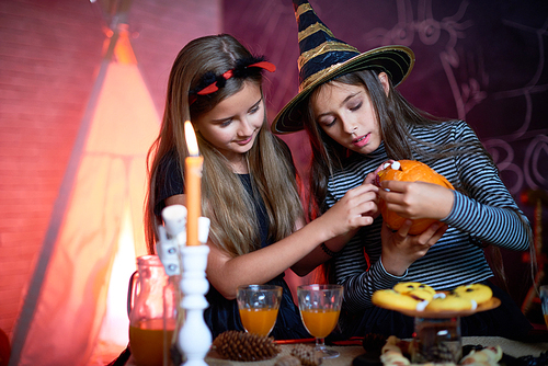 Portrait of two little girls wearing Halloween costumes making carved pumpkin in decorated room