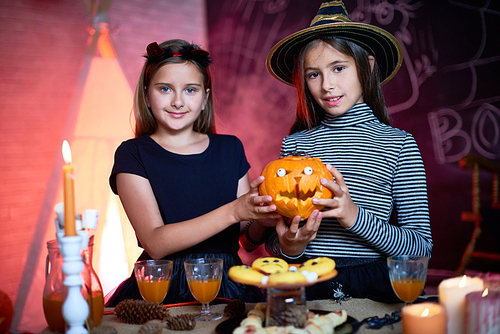 Smiling pretty friends dressed as witches holding carved pumpkin with eyeballs and  near candy bar at Halloween party