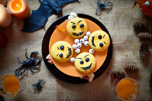 High angle view of creative Halloween cake in orange and yellow colors decorated emoticon cookies and eyeball candies placed on burlap with drinks in glasses, cones toy spiders and bat