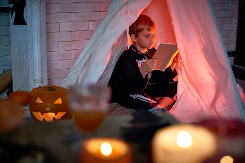 Concentrated cute boy dressed as skeleton sitting inside of teepee and reading interesting book alone in dark room during Halloween night