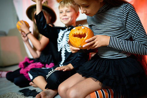 Closeup portrait of three children wearing Halloween costumes  sitting on floor in decorated room, focus on girl holding small carved pumpkin