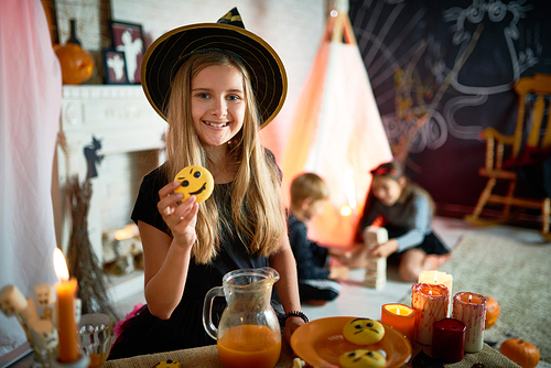 Portrait of pretty little girl dressed in Halloween costume posing with vampire cookie in decorated room, other children playing in background