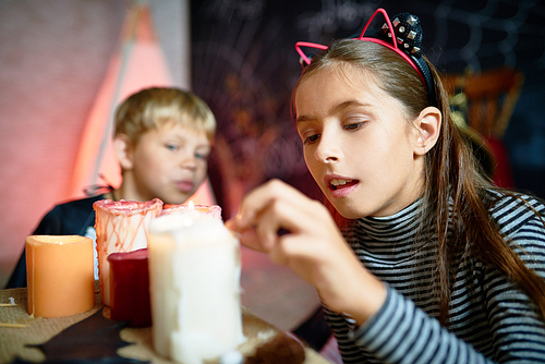 Portrait of pretty little girl lighting up candles on Halloween in decorated room, little boy wearing costume in background