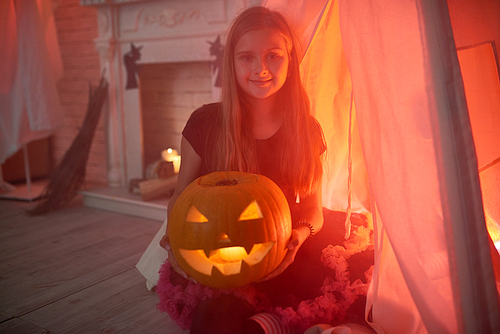 Portrait of pretty teenage girl holding pumpkin lantern smiling to camera in decorated room for Halloween