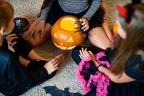 Above view of three children wearing Halloween costumes playing magic ritual with pumpkin lantern sitting on floor in circle