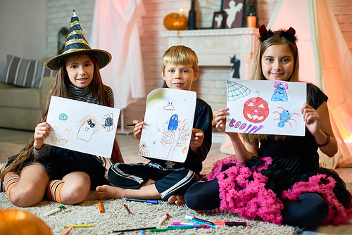 Positive cute friends in Halloween costumes sitting on carpet with splattering markers and showing creative pictures with Halloween characters to camera at home party