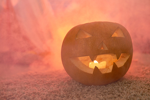 Close-up of mysterious jack-o-lantern with burning candle placed on carpet in red smoke