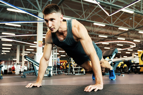 Portrait of handsome young man with muscular arms doing push ups on floor in modern gym