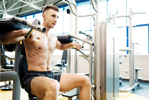 Portrait of handsome young man with bare chest pumping arm muscles doing exercises on machines in gym