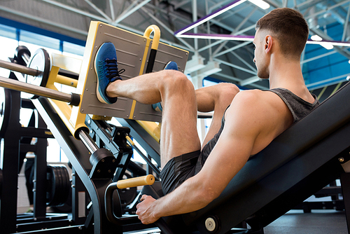 Rear view of muscular sportsman doing leg exercises using machine in modern gym