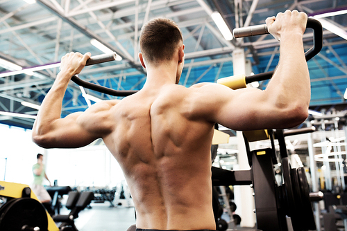 Back view portrait of handsome young man with bare chest pumping  muscles doing exercises on machines in gym