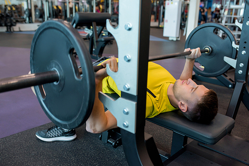 Portrait of young man doing weightlifting exercises with heavy barbell on bench during workout in modern gym