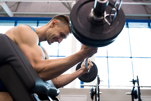 Side view portrait of handsome young man lifting barbell pumping arm muscles during strength training in modern gym, copy space