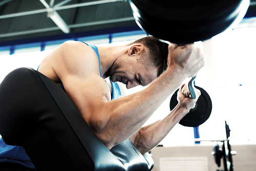 Side view portrait of strong man pumping arm muscles doing exercises with heavy barbell during workout in modern gym
