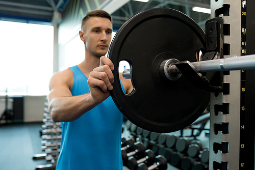 Portrait of handsome young man setting up barbell putting heavy plates on it during workout in modern gym