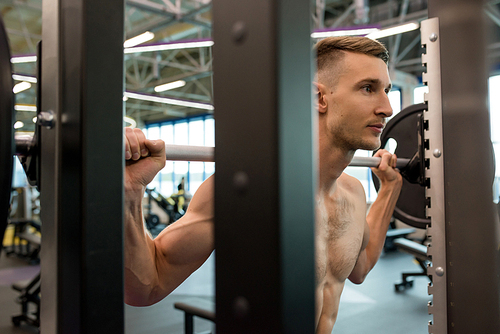 Portrait of handsome young sportsman with bare chest listing heavy barbell in gym, shot from behind weigh stand