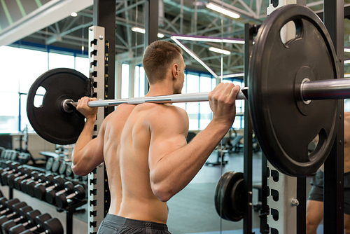 Rear view portrait of handsome young man with bare muscular back lifting heavy barbell during workout in modern gym