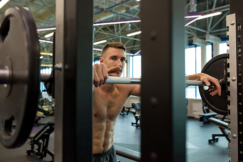 Portrait of shirtless young man lifting barbell from stand during weightlifting training in modern gym