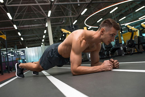 Portrait of young muscular man with bare chest doing plank exercise on floor during workout in modern gym