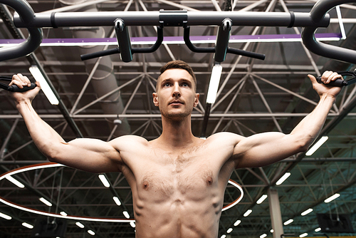 Low angle portrait of determined young man with bare chest pumping arm muscles working out on machines in gym