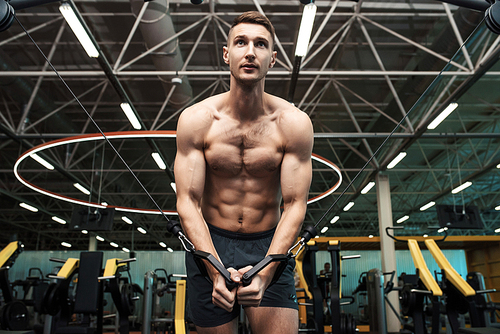 Low angle portrait of determined young man with bare chest pumping muscles working out on machines in gym  in dramatic light