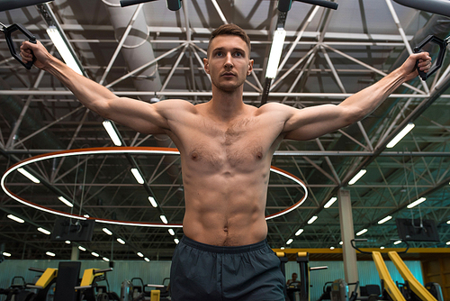 Low angle portrait of motivated young man with bare chest working out using machines  in modern gym against background on metal ceiling