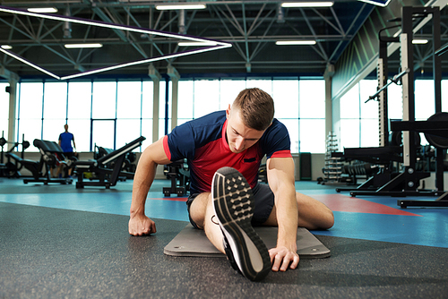 Portrait of young man stretching legs on floor while warming up for workout in modern gym