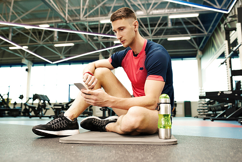 Portrait of handsome young man sitting on floor in gym using smartphone taking break from workout to relax