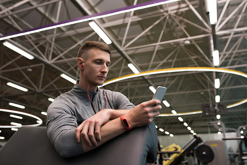 Portrait of handsome sportsman wearing fitness bracelet using smartphone listening to music in modern gym taking break from workout
