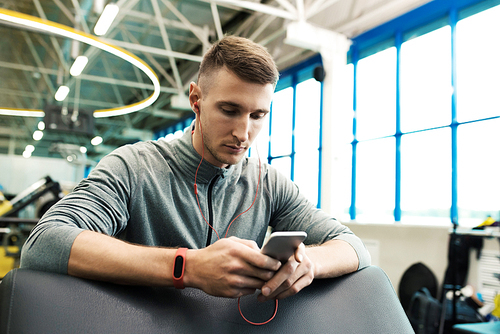 Portrait of handsome young man using smartphone listening to music in modern gym taking break from workout