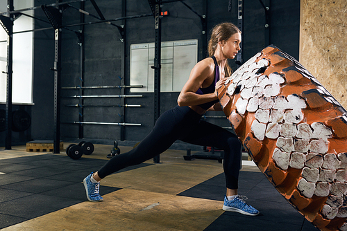Side view portrait of strong young woman flipping heavy tire during crossfit workout in modern gym