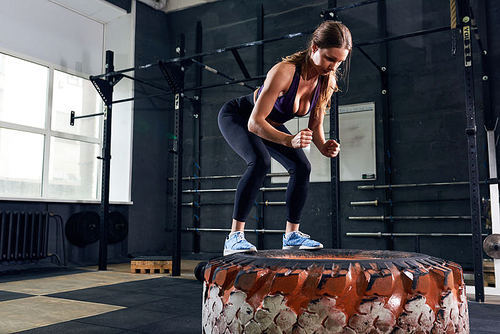 Portrait of strong young woman doing box jumps on tire during intense workout in modern gym