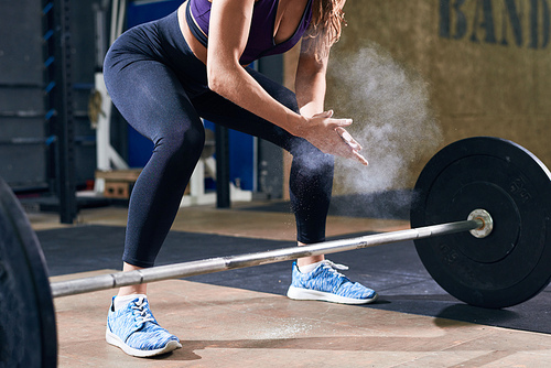 Low section portrait of unrecognizable young woman rubbing her hands with talk puffing small cloud preparing to lift heavy barbell during crossfit workout in modern gym.