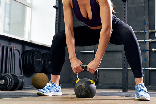 Low section portrait of unrecognizable young woman weightlifting, squatting with kettlebell during crossfit workout in modern gym