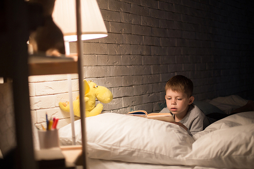 Portrait of cute little boy reading fairytales in bed under lamp light, propping book on pillow in dark room at night, copy space