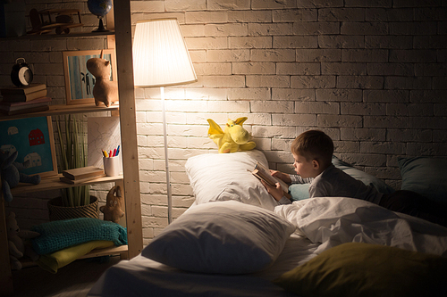 Side view portrait of cute little boy reading fairytales in bed under lamp light, propping book on pillow in dark room at night, copy space