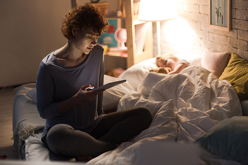 Full length portrait of young woman sitting on bed reading bedtime story to child sleeping next to her in dim lamplight, copy space