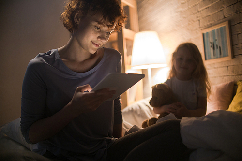 Portrait of young woman reading bedtime story to child sitting on bed and smiling in dim lamplight