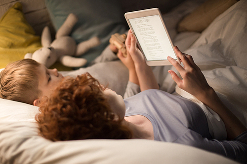 Portrait of mother and son reading book together from tablet lying in bed before sleep, enjoying stories in dim lamplight