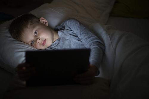 Portrait of little boy using computer in bed, face lit by screen light playing games late at night in dark