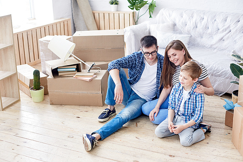 Cheerful young family sitting on floor of new apartment and sharing impressions with each other, pile of moving boxes on background