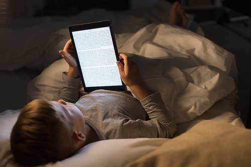 High angle portrait of little boy reading book in bed using e-reader at night, face lit by white screen light