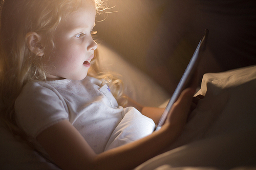 Side view portrait of adorable little girl looking amazed with mouth open using digital tablet lying in bed at night, face lit by screen light like magic, copy space