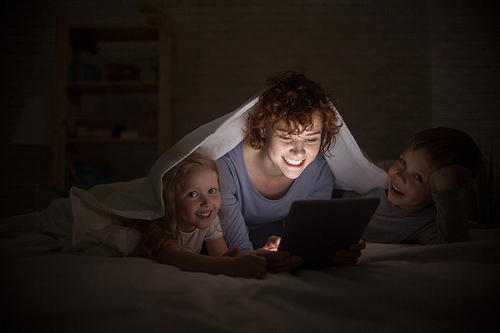 Portrait of happy young mother with two children reading stories in bed, hiding under blanket and using e-book
