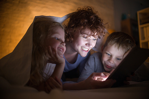 Portrait of happy young mother with two children reading stories in bed, hiding under blanket and using tablet