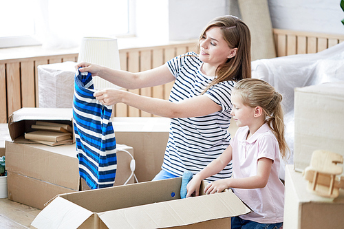 Attractive young mother and pretty little daughter gathered together at living room and packing cardboard box in order to move into new apartment