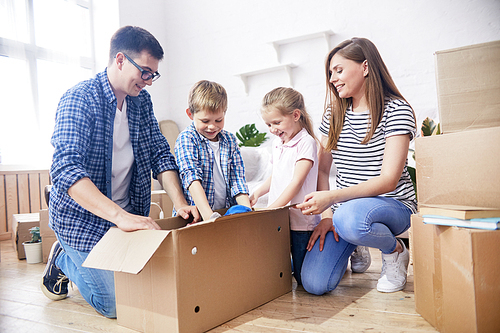 Cheerful young family gathered together at spacious living room of new apartment and enthusiastically unpacking moving boxes with their stuff